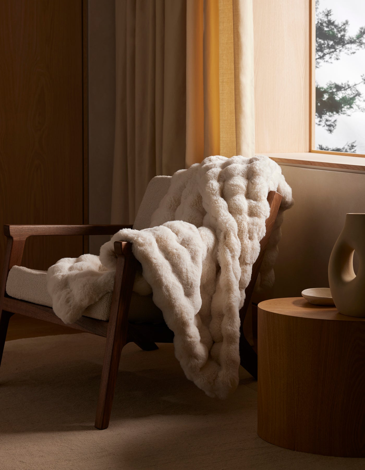 The Cozy Earth Bubble Cuddle Blanket, in cream, is draped over a wooden armchair next to a round wooden side table with beige curtains and soft natural light filtering through the window.|Color:Creme