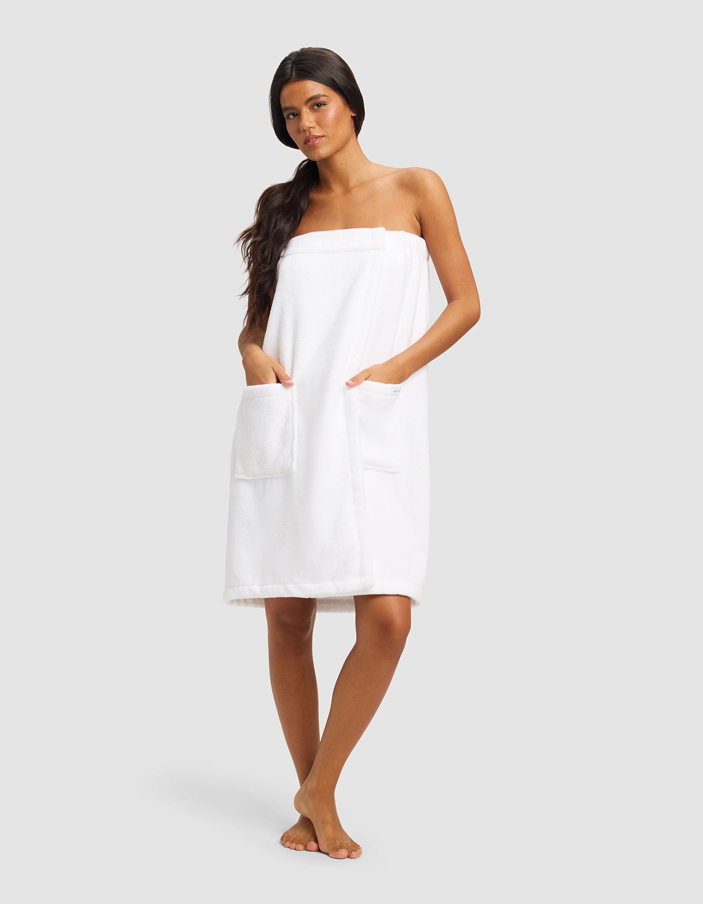 A woman with long dark hair stands barefoot against a plain background, wearing Cozy Earth's Luxe Bath Wrap featuring two large front pockets.|Color:White