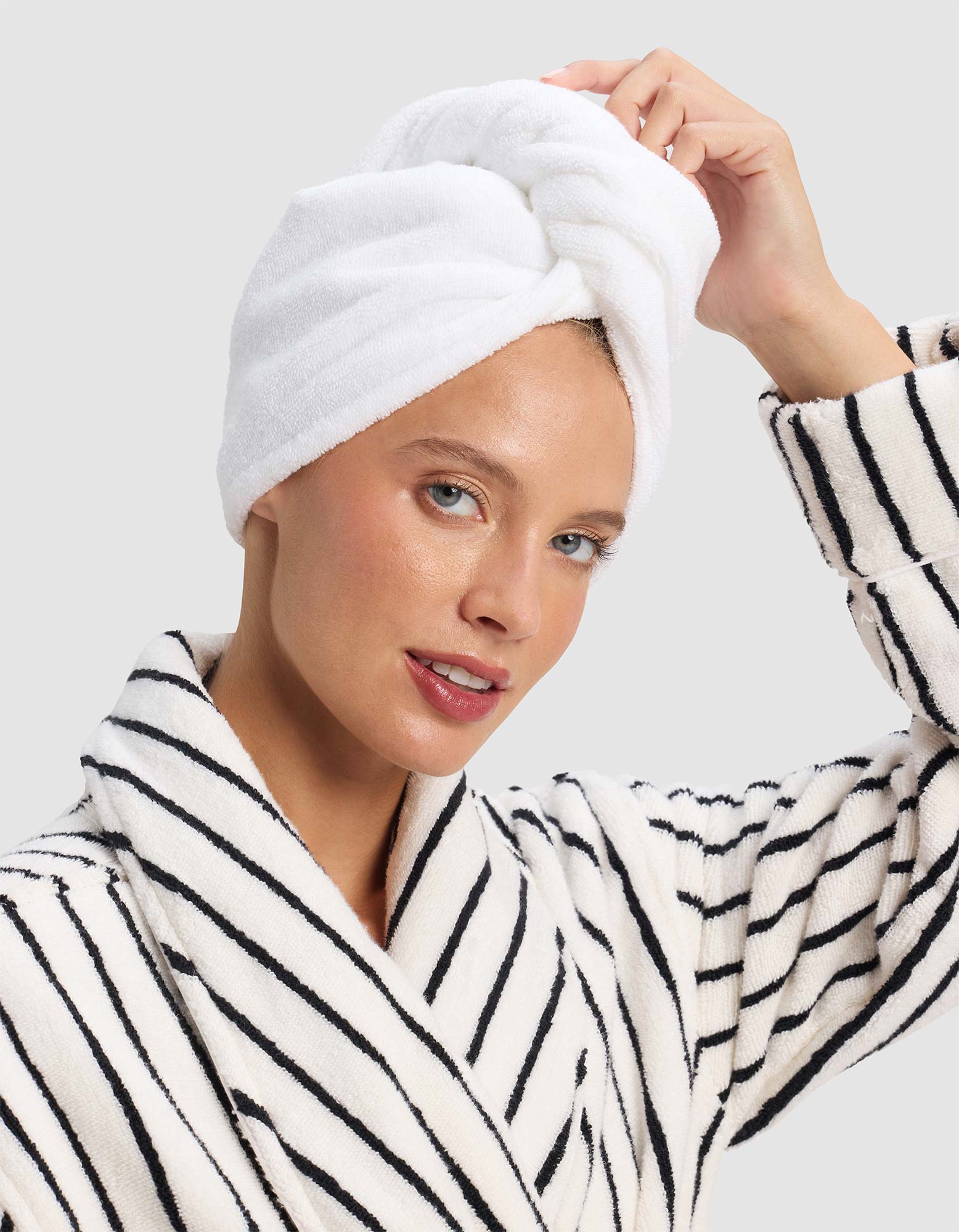 A woman smiles softly, touching the Cozy Earth Luxe Hair Towel wrapped around her head, while wearing a white bathrobe with black stripes against a plain light background. |Color:White