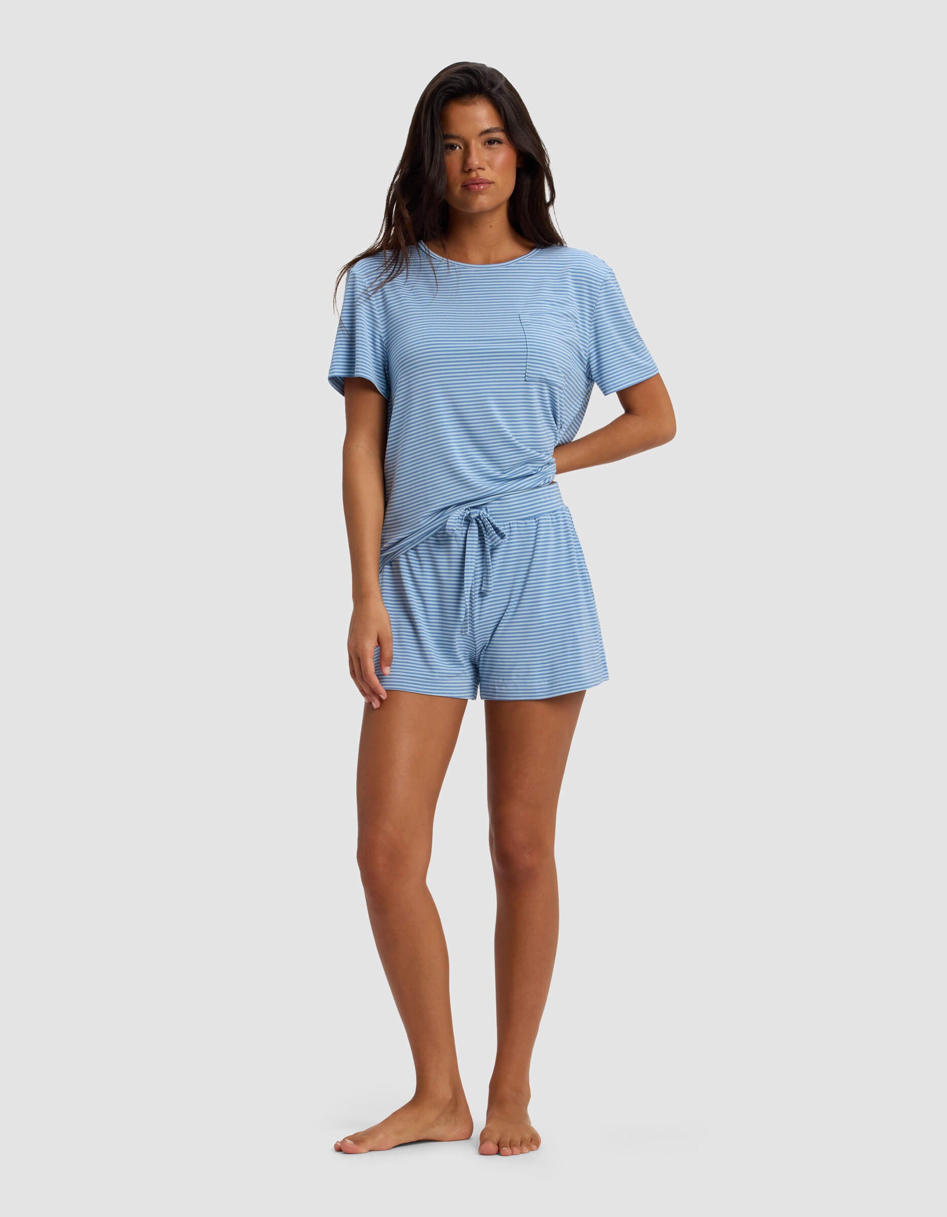 A woman stands barefoot against a plain background, wearing the Cozy Earth Women's Bamboo Stretch-Knit Lounge Tee & Short Set in light blue with short sleeves, subtle stripes, a chest pocket, and a waist tie.|Color:Sailor Mini Stripe