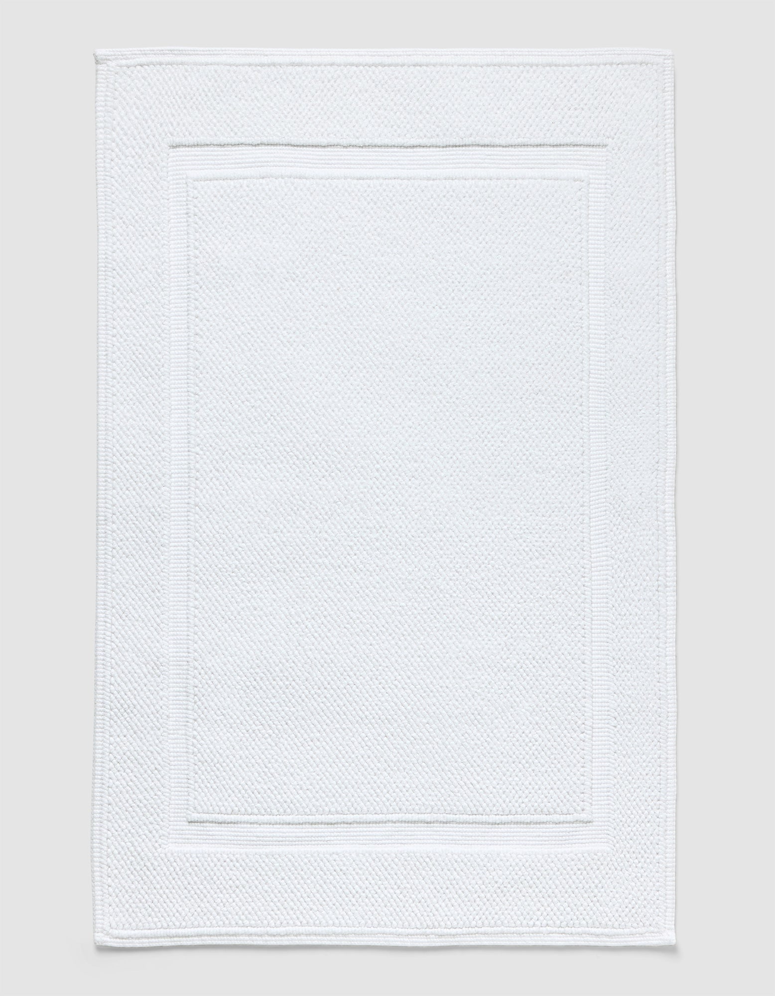 The Cozy Earth Classic Bath Mat in white features a textured surface with a subtle border pattern and is shown flat against a neutral background.|Color:White