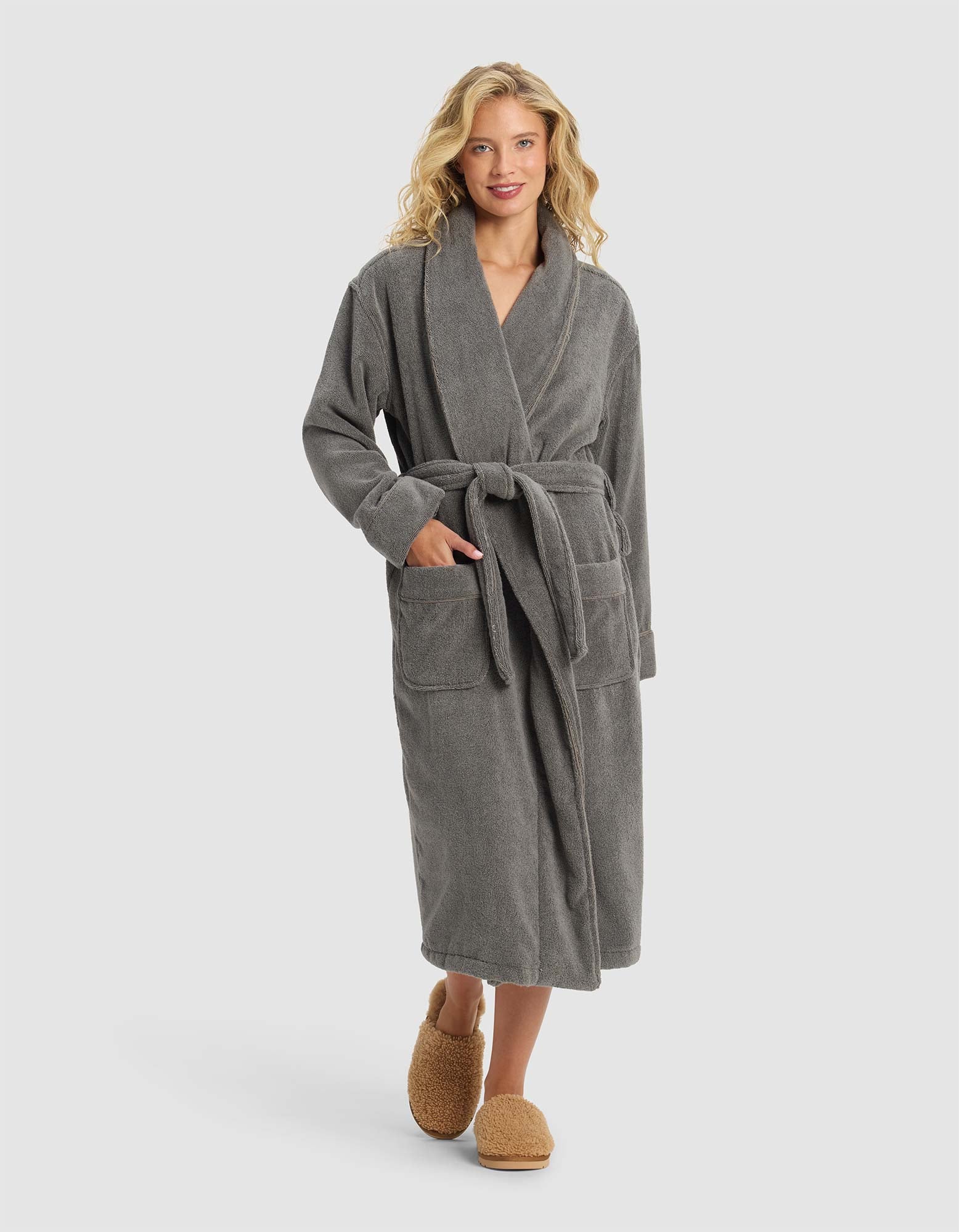 A woman with wavy blonde hair smiles while wearing the Cozy Earth Luxe Bath Robe in gray, featuring a belted waist and large pockets, paired with brown slippers, against a plain light background.|Color:Charcoal