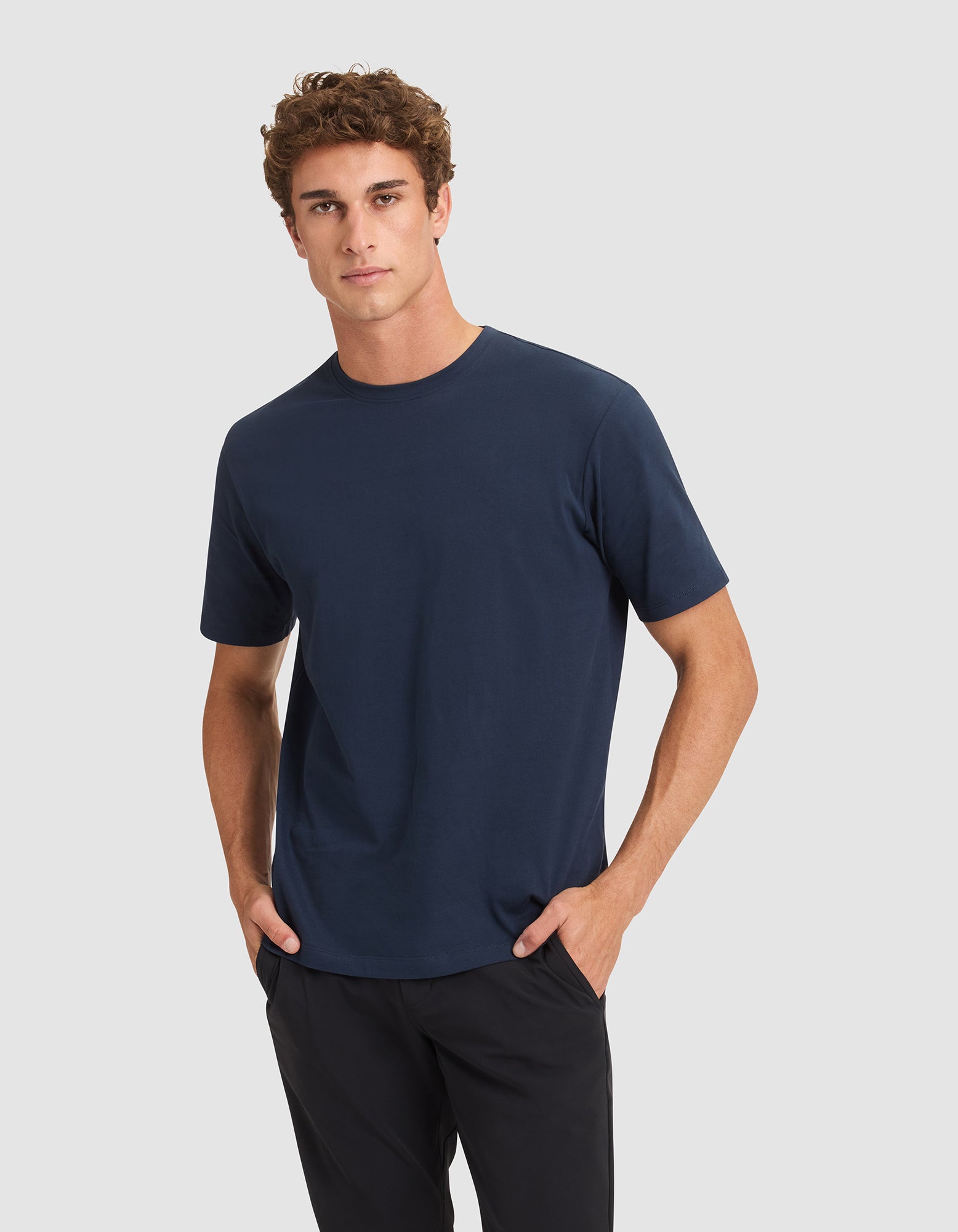A young man with curly brown hair stands with his hands in his pockets against a light gray background, wearing the Cozy Earth Men's All Day Tee in navy blue and black pants. |Color:Eclipse