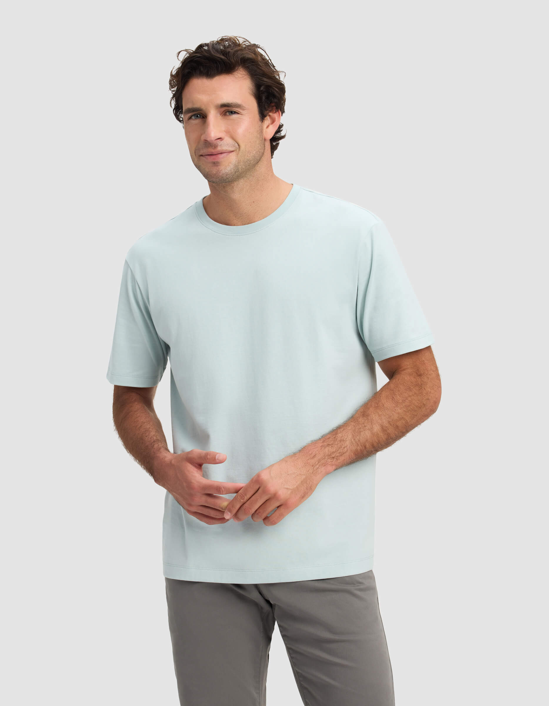 A man with short brown hair is wearing the Cozy Earth Men's All Day Tee in light blue and gray pants, standing against a plain light gray background and smiling slightly.|Color:Tide
