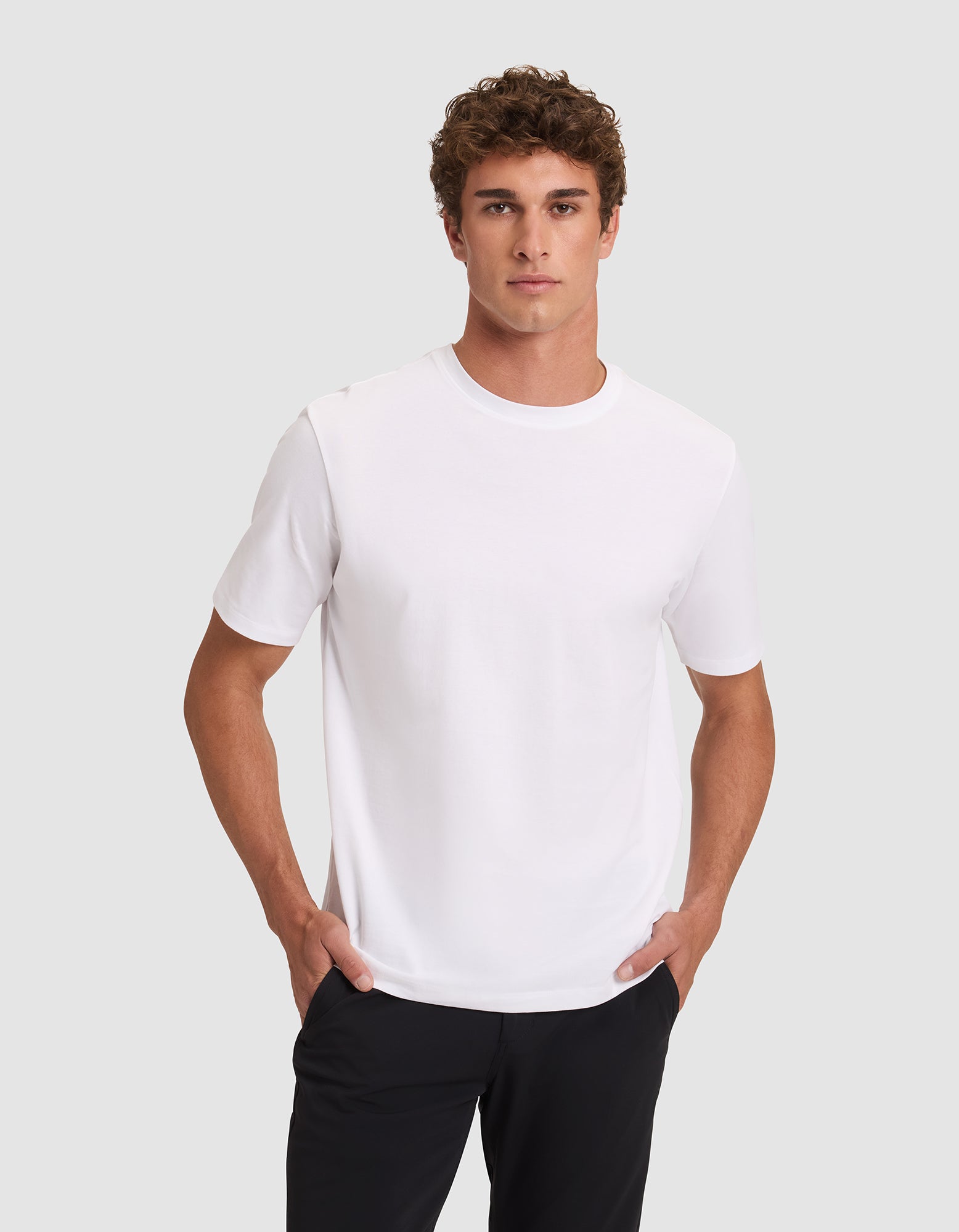 A man with short curly hair wears the Men's All Day Tee by Cozy Earth in white, paired with black pants, standing against a light gray background with his hands in his pockets and looking at the camera neutrally. |Color:White