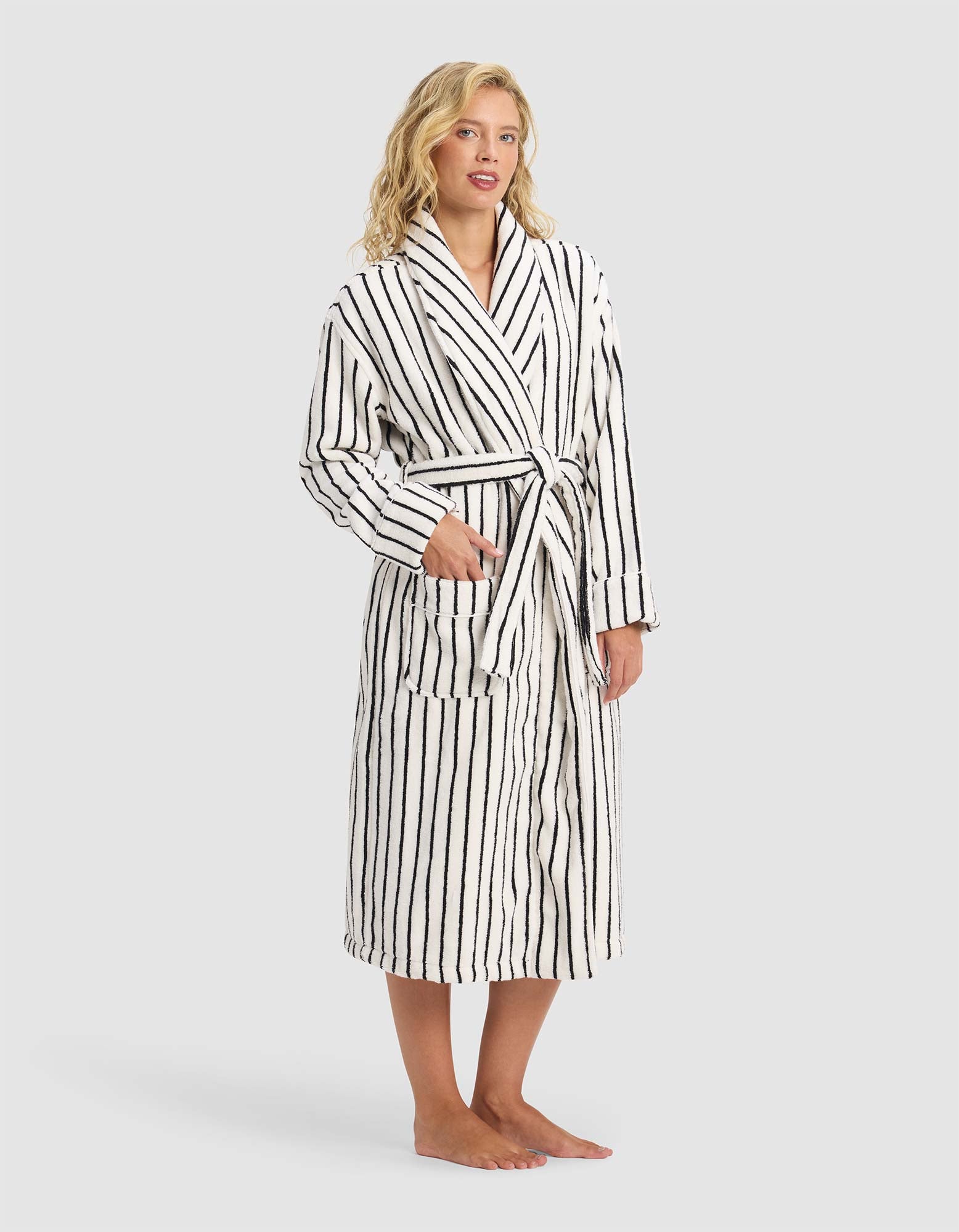 A person stands barefoot in the Cozy Earth Striped Luxe Bath Robe, white with black vertical stripes and a tie waist, hands partially in pockets, against a plain light gray background.|Color:Black Stripe